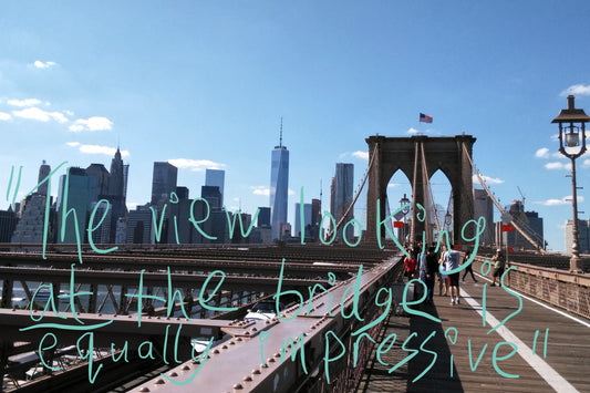 View of the New York City skyline with the Freedom Tower in shot, from the perspective of being in the pedestrian area of the Brooklyn Bridge. Graffiti overlay featuring a quote from the article: "The view looking at the bridge is equally impressive"