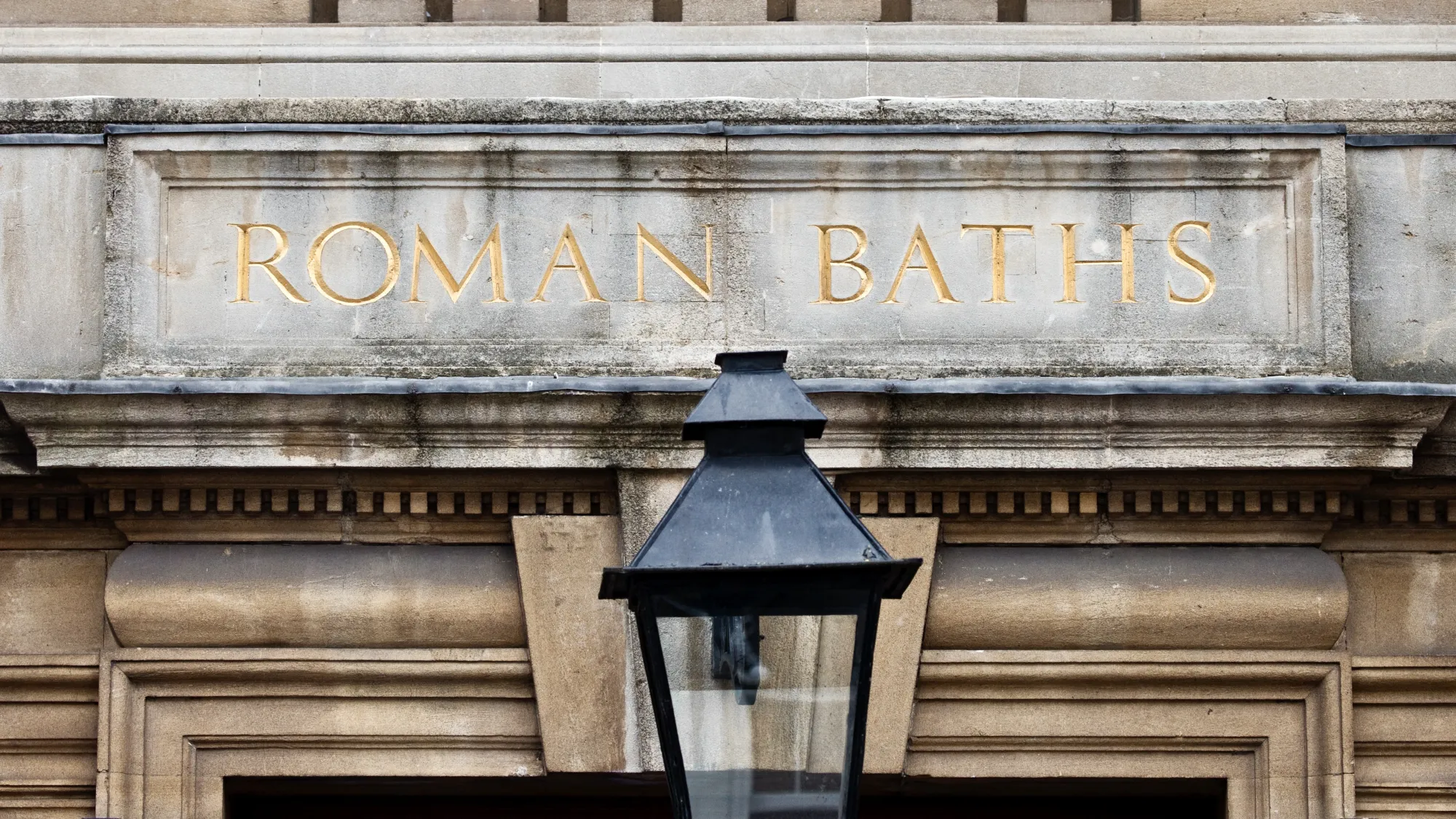 The top of a stone doorway with the words 'Roman Baths' carved into it. Gold colouring is inlaid into the carved wording