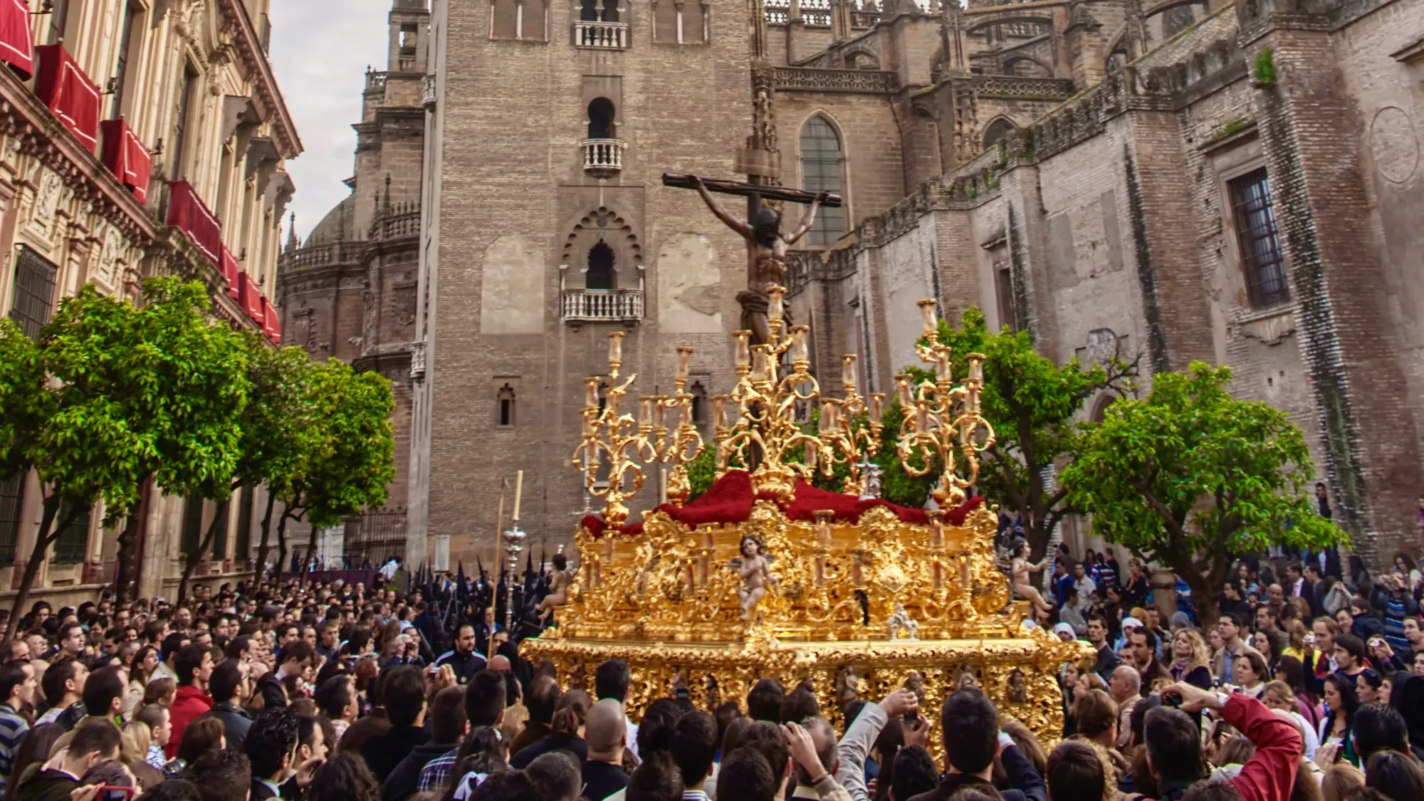 The Semana Santa event in Sevilla, otherwise known as Holy Week. There is a gold float with religious iconography, held aloft by crowds of people. The streets are lined by oranges trees.