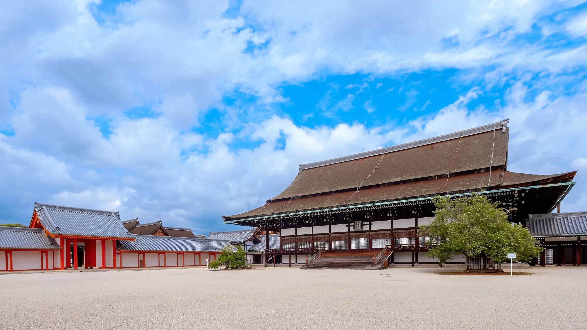 The Kyoto Imperial Palace. This was the residence of the Emperor of Japan, until 1868 when the Meiji period began and the Emperor moved to Tokyo. The sky is blue, with some clouds. There is a wide, clean expanse of white gravel before the traditional Japanese Palace.