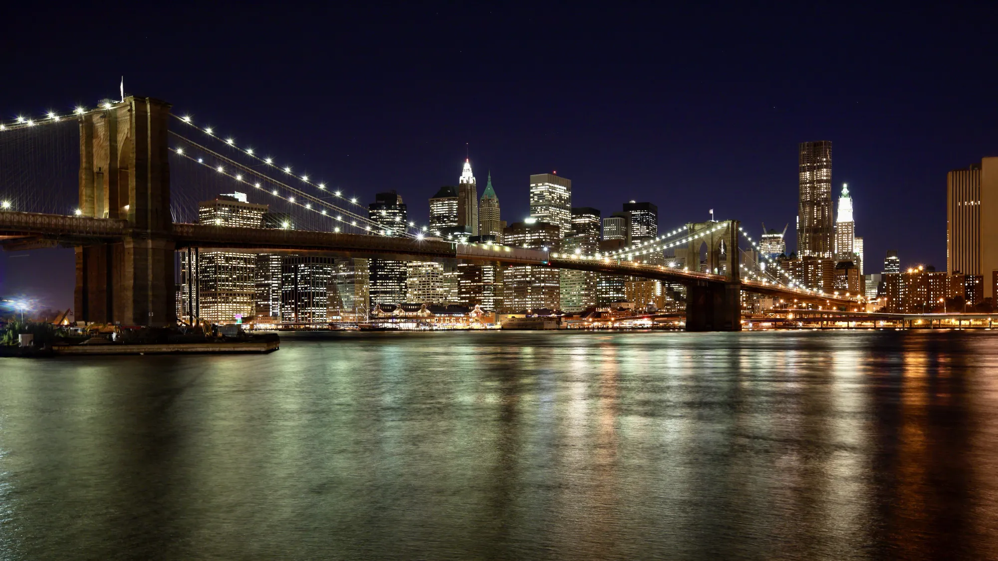 New York's Brooklyn Bridge lit up at night. The backdrop features the New York skyline covered in its iconic twinkling lights