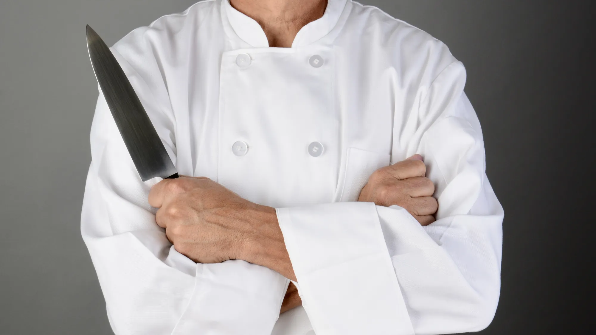 A shot of a chef's folded arms. The chef is wearing chef whites and has a steel kitchen knife in their hand