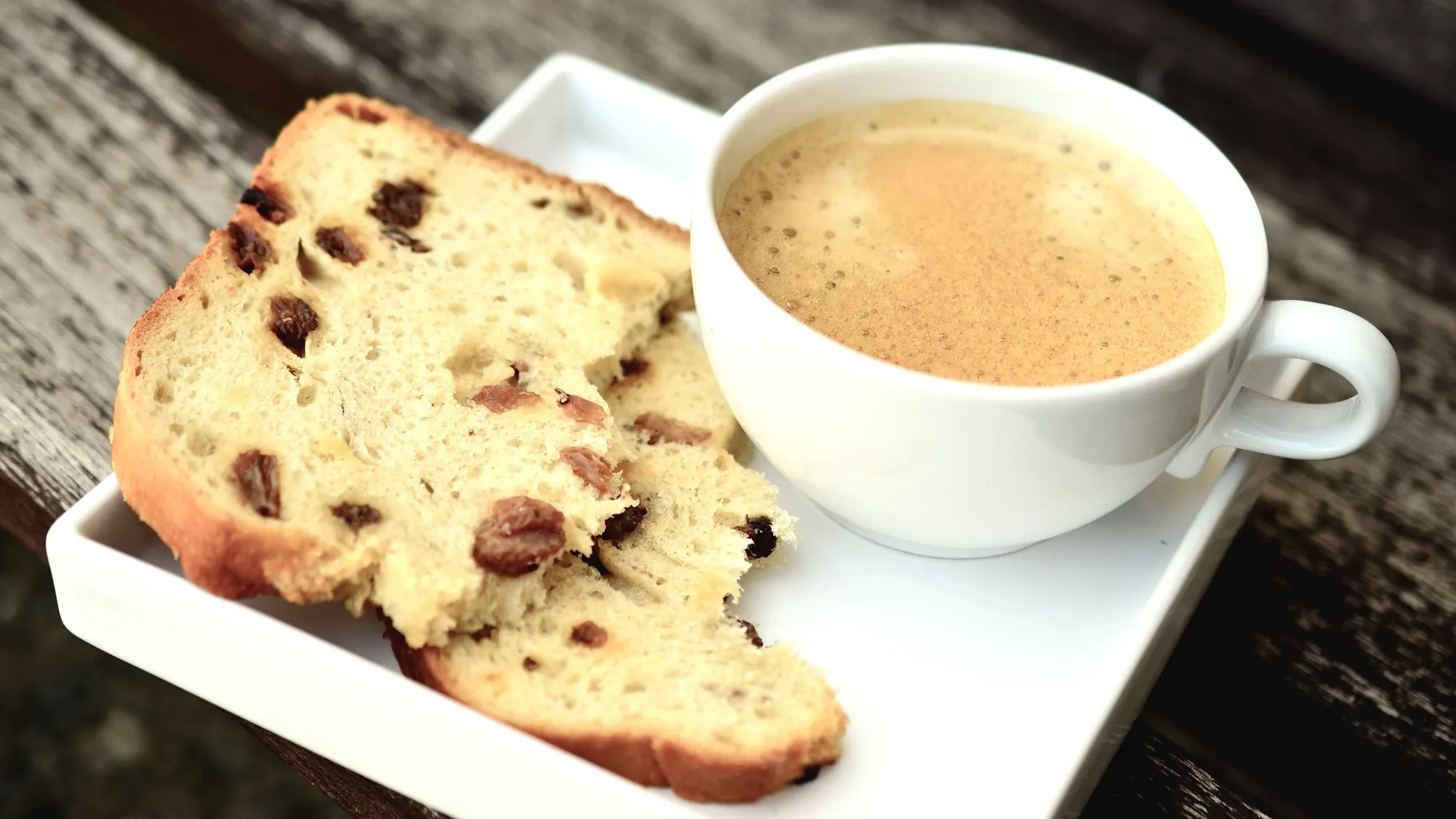 A cup of coffee with a coffee and cream coloured froth on top. The cup of coffee is sat on a small plate. The small plate also has a slice of fruit bread on it. The fruit bread is broken in half and one piece is stacked atop of the other