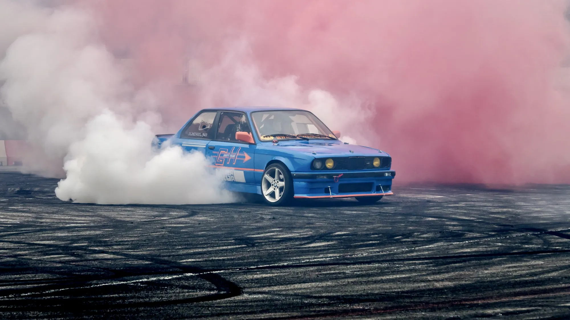 An action shot of a blue BMW drift car surrounded by pink and white smoke. The smoke is the result of the car having drifted around the tarmac area in the foreground. The tarmac area is covered in black tyre marks