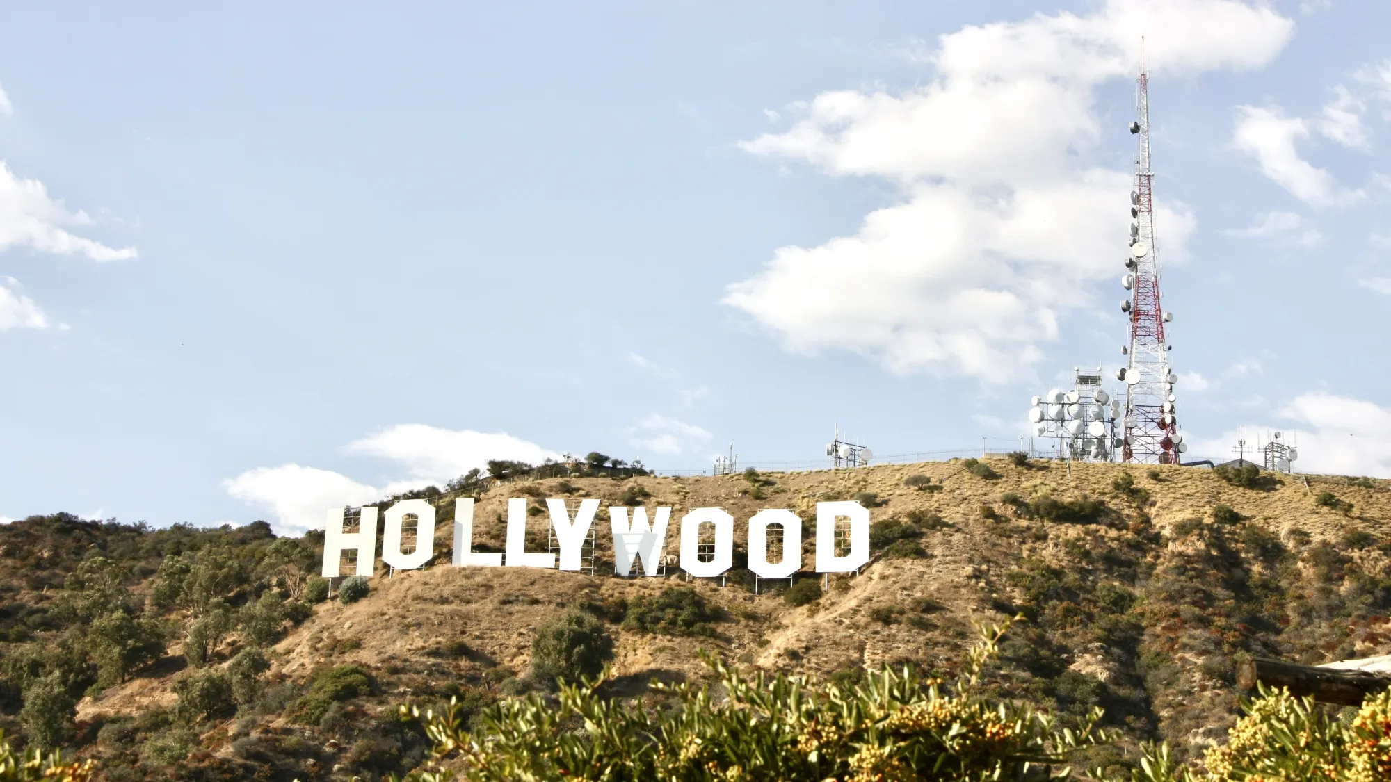 The Hollywood Sign, on the side of the Hollywood hills. A pale blue sky can be seen in the background and the Hollywood radio mast is to the right of the sign