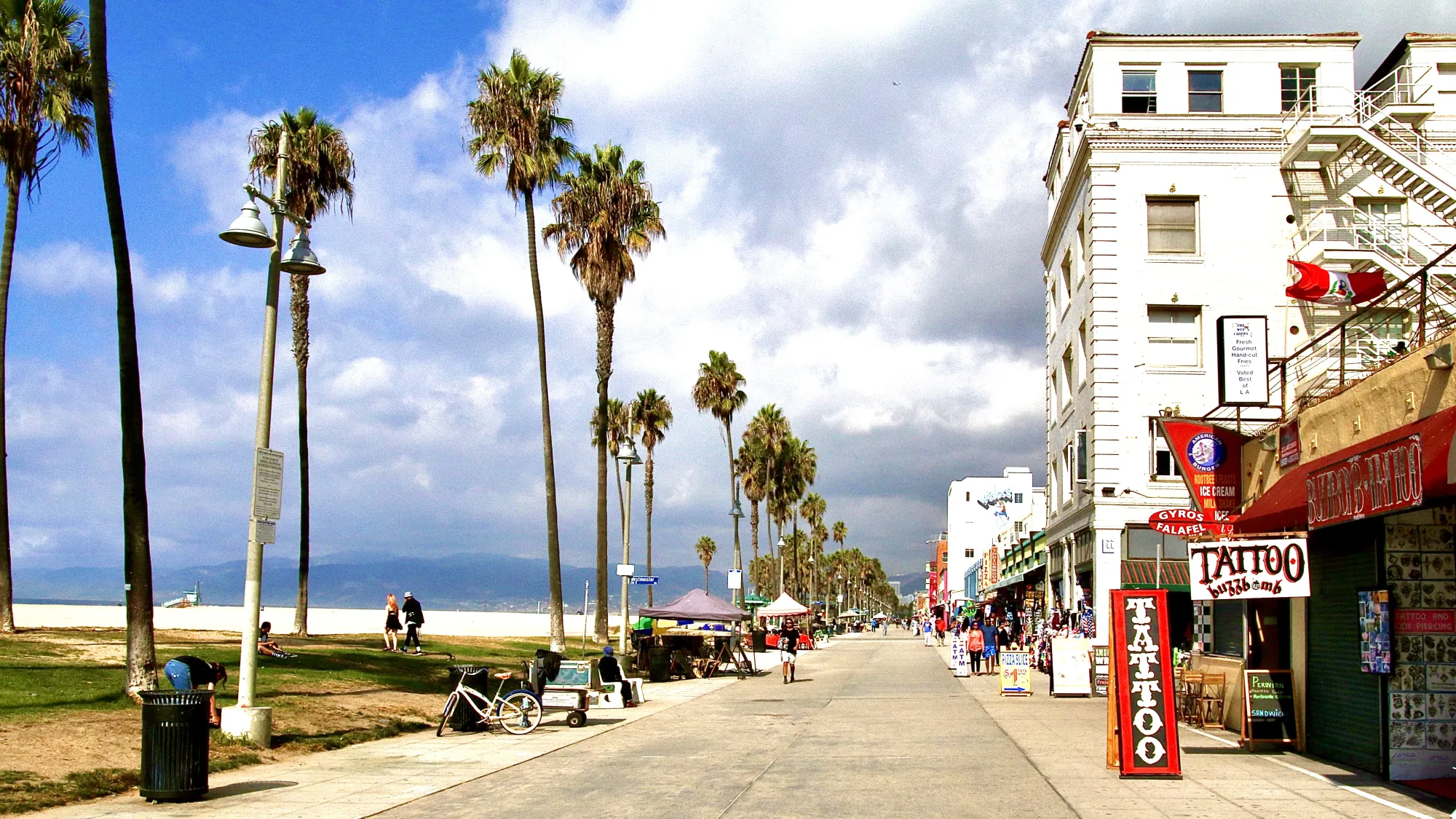 Los Angeles' Venice Beach boardwalk. The boardwalk is has a line of palm trees and the beach to the left and shop frontages to the right. The shopfront closest to the front of the shot is a largely red coloured tattoo shop