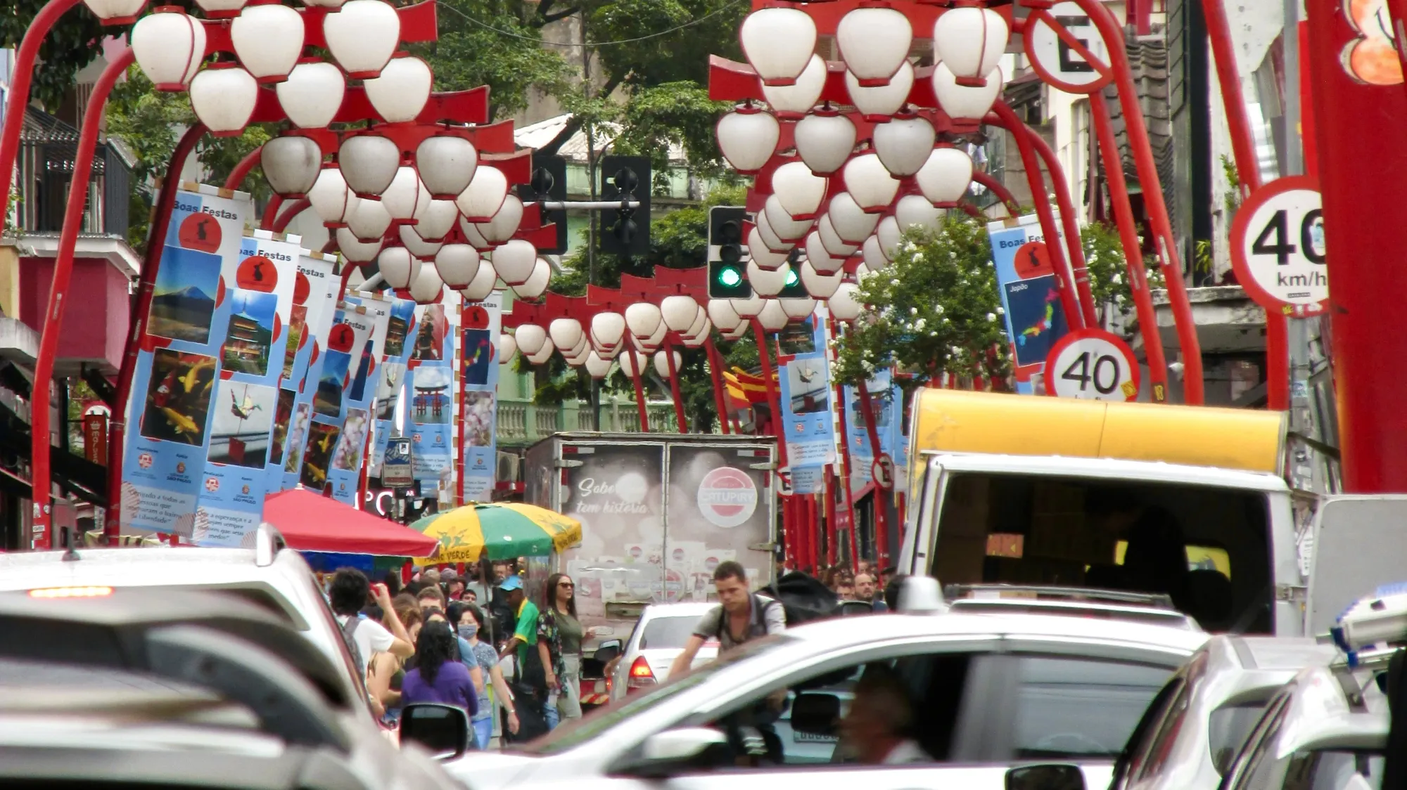 A very busy area in the Liberdade area of São Paulo. In the shot we can see cars, cyclists and market stalls. The focus of the image seems to be the red lampposts, which have white lantern style lights at the ends of them. There are people walking through the market area around the traffic. On the lampposts, there are posters displaying information about Japan and Japanese customs.