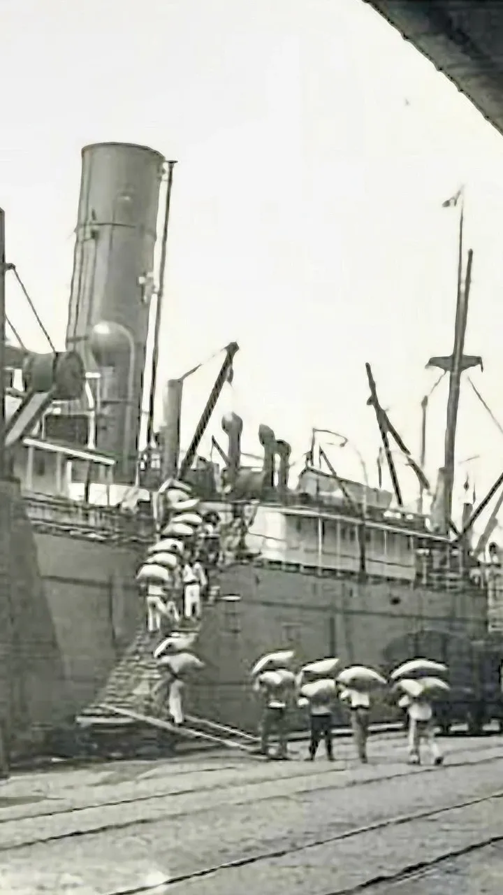 An old 1895 black and white photograph of people loading heavy sacks onto a steam ship. The ship is docked at Santos Port. It is likely the heavy sacks are filled with crops for sale overseas - probably coffee beans.