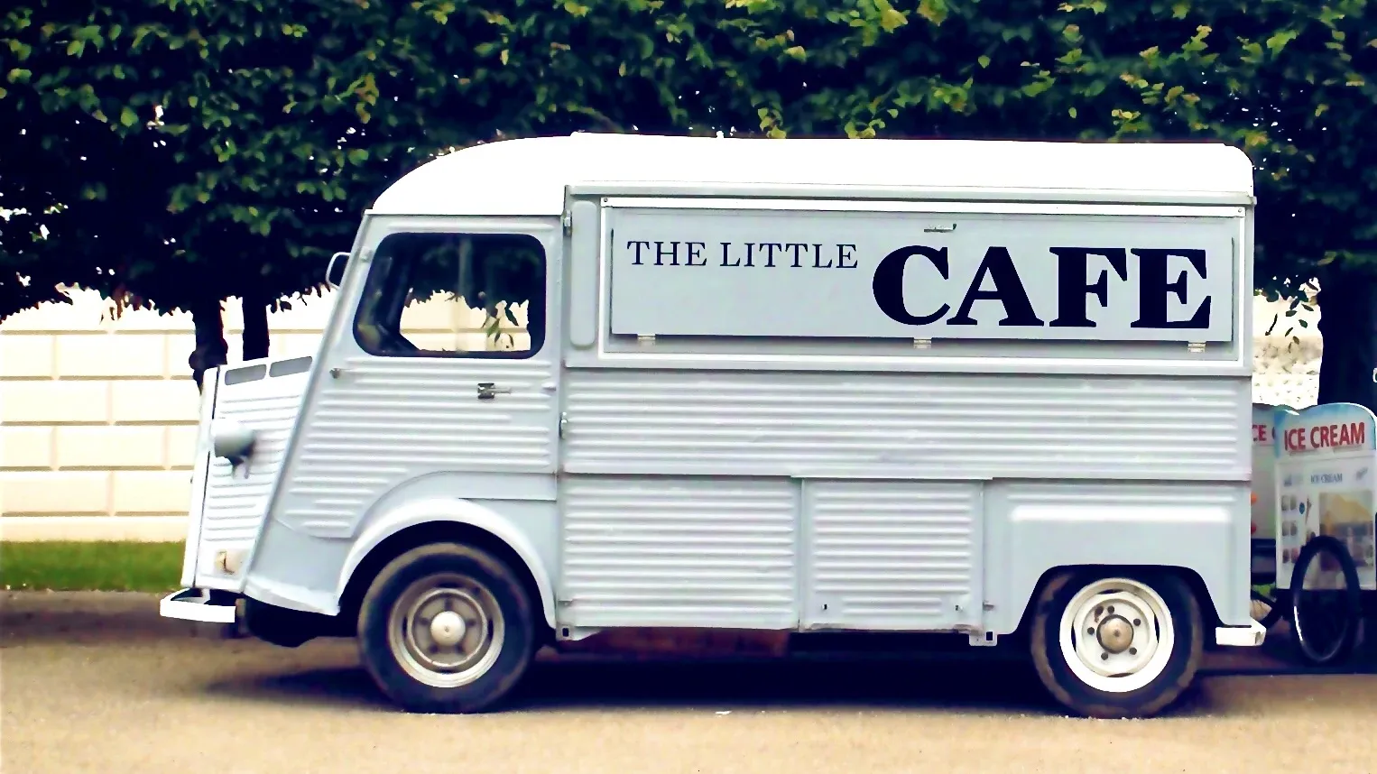 An old pale blue van with a serving hatch to sell food from. The hatch is closed and written on it are the words "The Little Cafe"