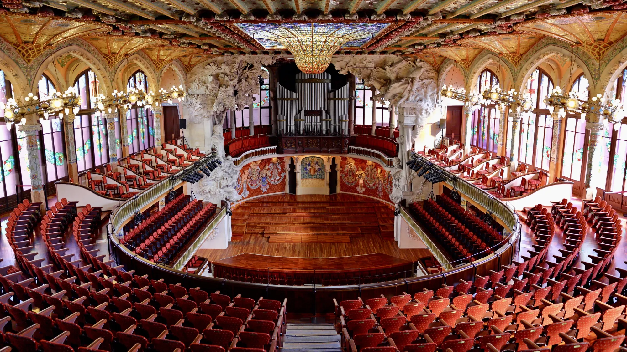 The inside of Barcelona's Palau de la Música Catalana. There is a large organ at the far end of the room and concert hall seating curved around the room. The centrepiece of this breathtaking piece of architecture is the 3-dimensional curved stained glass ceiling, and the ornate carved stonework