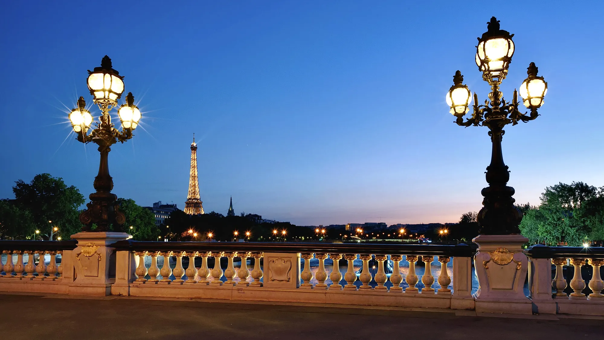 A bridge in Paris with two famous Parisian Arc Lamp style street lights, also known as Yablochkov Candles. In the background we can see a blue sky and a pink sunset with the Eiffel Tower alight with golden coloured lights