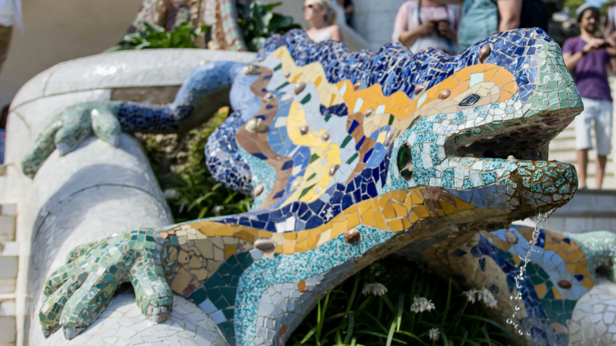 A stone salamander lizard covered in colourful mosaic tiles. The lizard is sitting between two flights of stairs at the entrance of Barcelona's Park Güell. A trickle of water is falling from the lizard's mouth. The lizard's name is El Drac