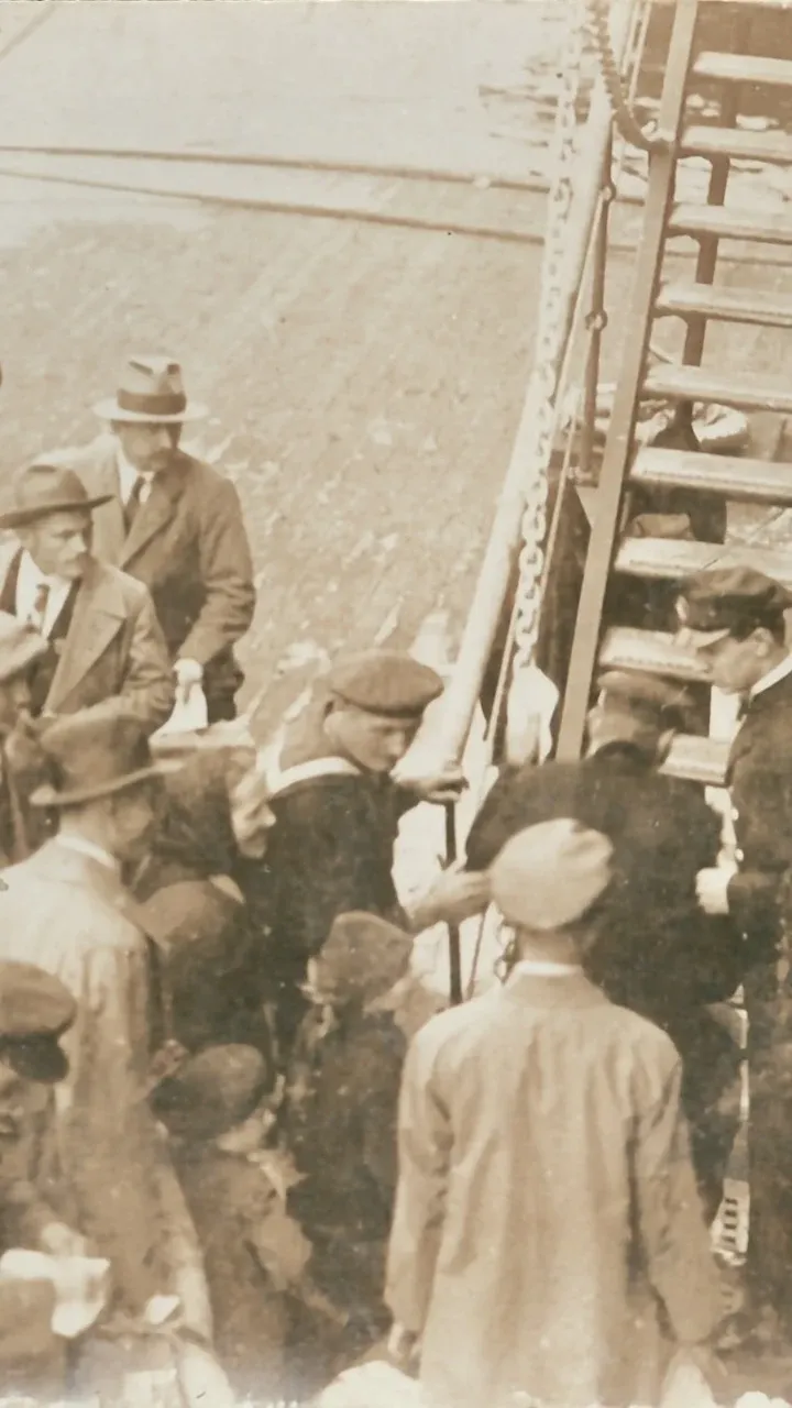An old 1898 sepia-tone photograph of men, women and children about to climb the stairs to board a ship bound for Brazil. The people are boarding in Italy, but are of mixed European origin. It is thought the photographer is Swiss.