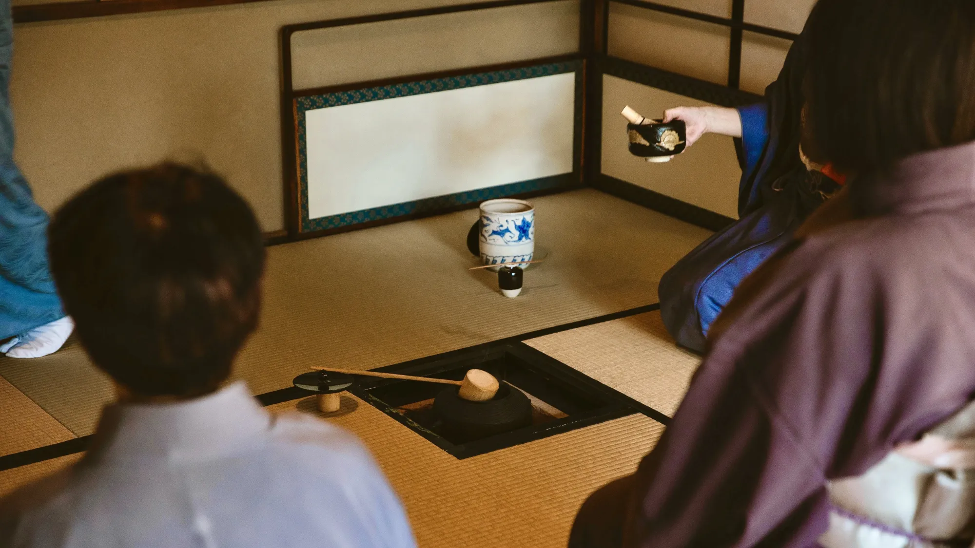 A traditional tea ceremony, which is taking place in a Kyoto tea room. The participants are sat on Tatami mats and the typical tea ceremony apparatus can be seen in the centre.