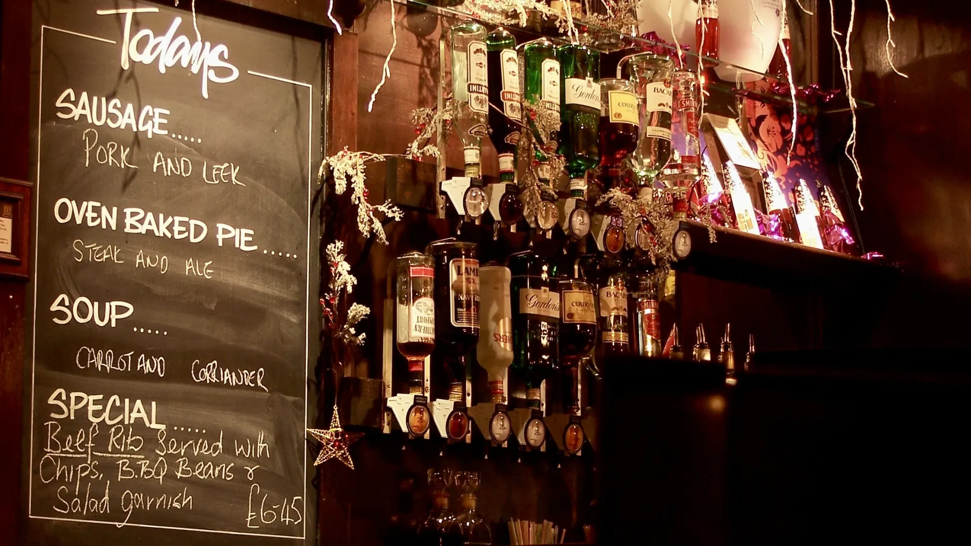 A menu board, next to some bottles of spirits in a traditional British pub. The menu board lists the day's specials, including "Pork and Leek Sausage", "Steak and Ale Oven Baked Pie", "Carrot and Coriander Soup", and a special meal which is "Beef Rib Served with Chips, BBQ Beans & Salad Garnish"