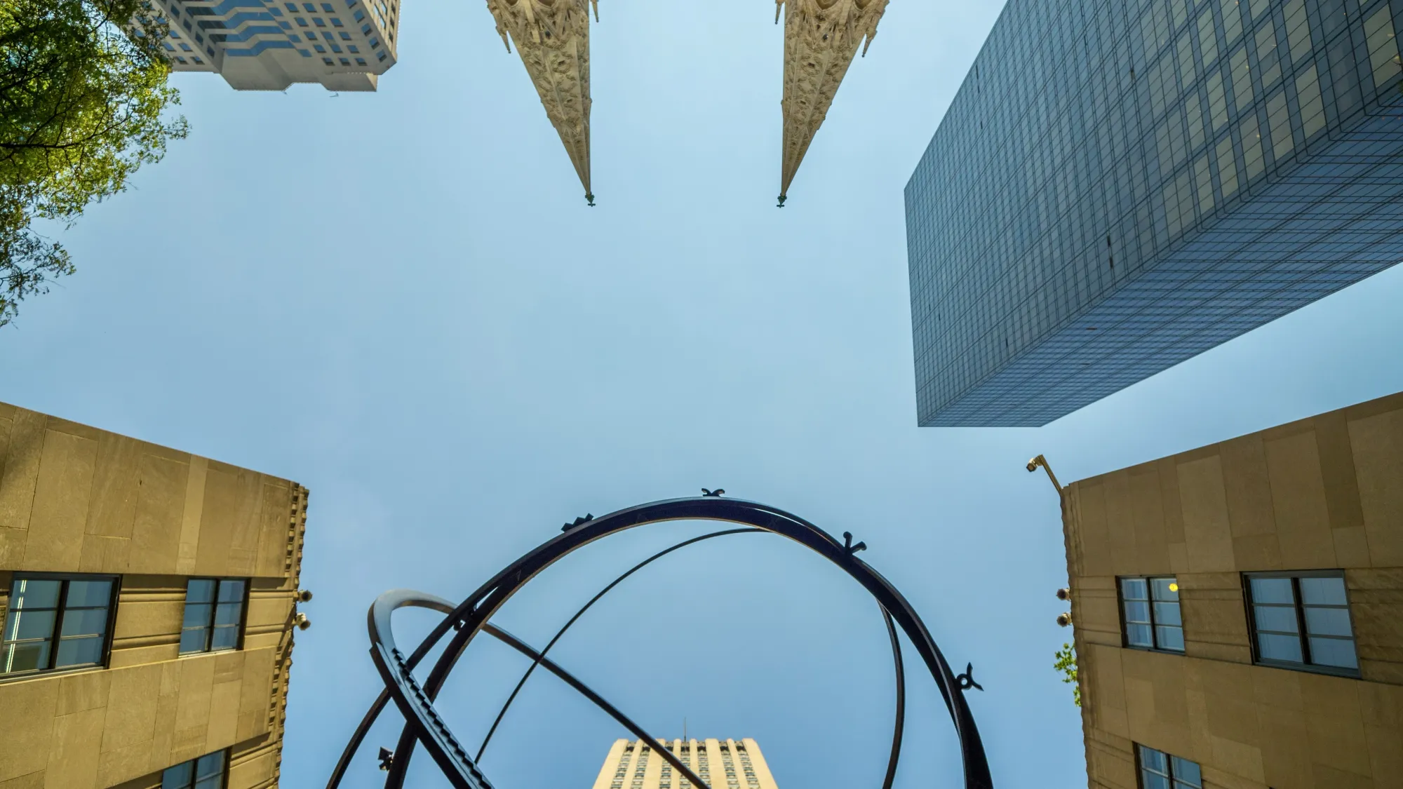 Looking skywards from the ground, at the the buildings in the Rockefeller Center. The blue sky fills the centre of the picture and some of the Rockefeller buildings surround the edges of the frame. At the bottom of scene we can see the sphere of the famous statue of the Greek God 'Atlas'