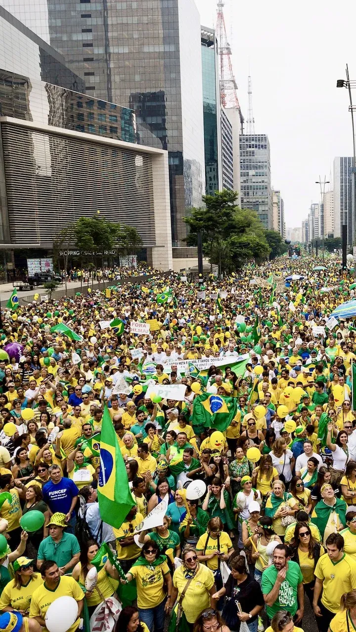 A São Paulo street full of people wearing the colours typically associated with Brazil. The street is filled with people width-ways across both sides of the road and length-ways stretching as far as the eye can see into the distance.