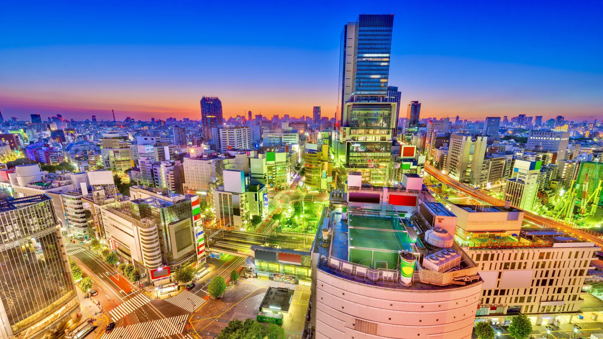 Tokyo cityscape with a stunning sunset in the background. The buildings in the scene are emitting a green hue as a result from the many lights in the area. The sights of note in the foreground are the Shibuya scramble crossing and - the main focus point - the Shibuya futsal park, which is a well-known rooftop football pitch