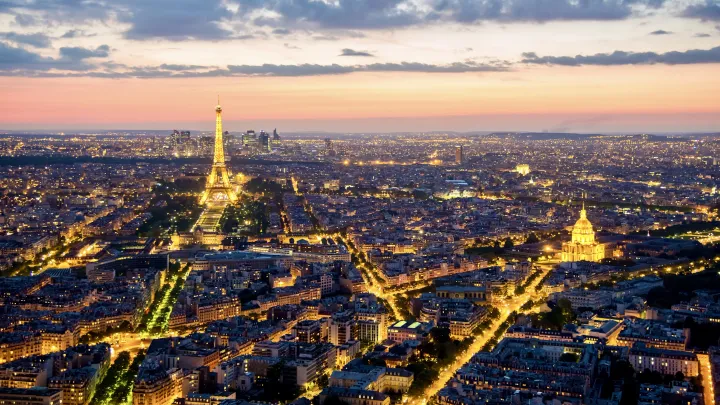 View of the Paris skyline with a dusk sky, the Eiffel Tower and Les Invalides are lit up by golden lights and the Arc de Triomphe can be seen in the background.