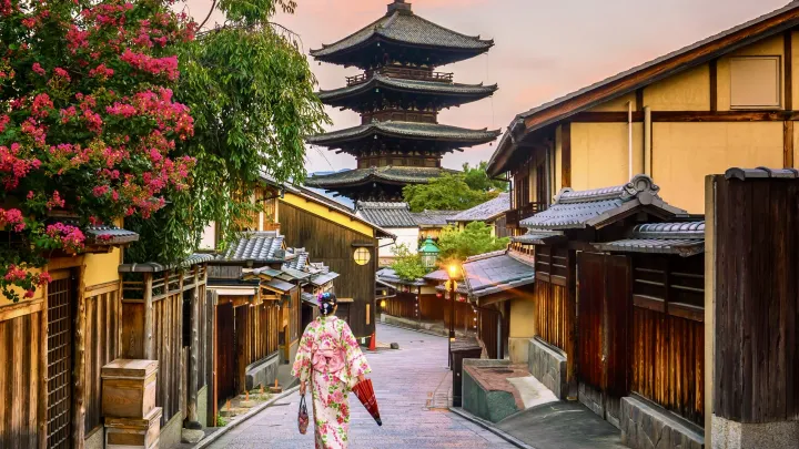 Street in Kyoto old town, pagoda in distance, soft pink sky. Traditionally dressed Japanese lady walking away from camera, holding red umbrella.