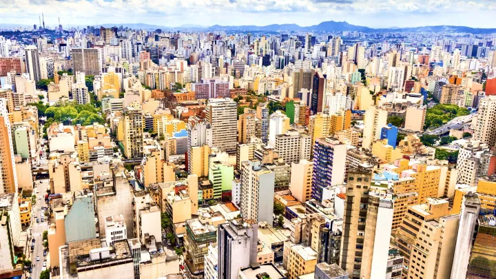 An aerial photo of São Paulo, buildings are densely packed. The skies are blue, with white clouds passing through.